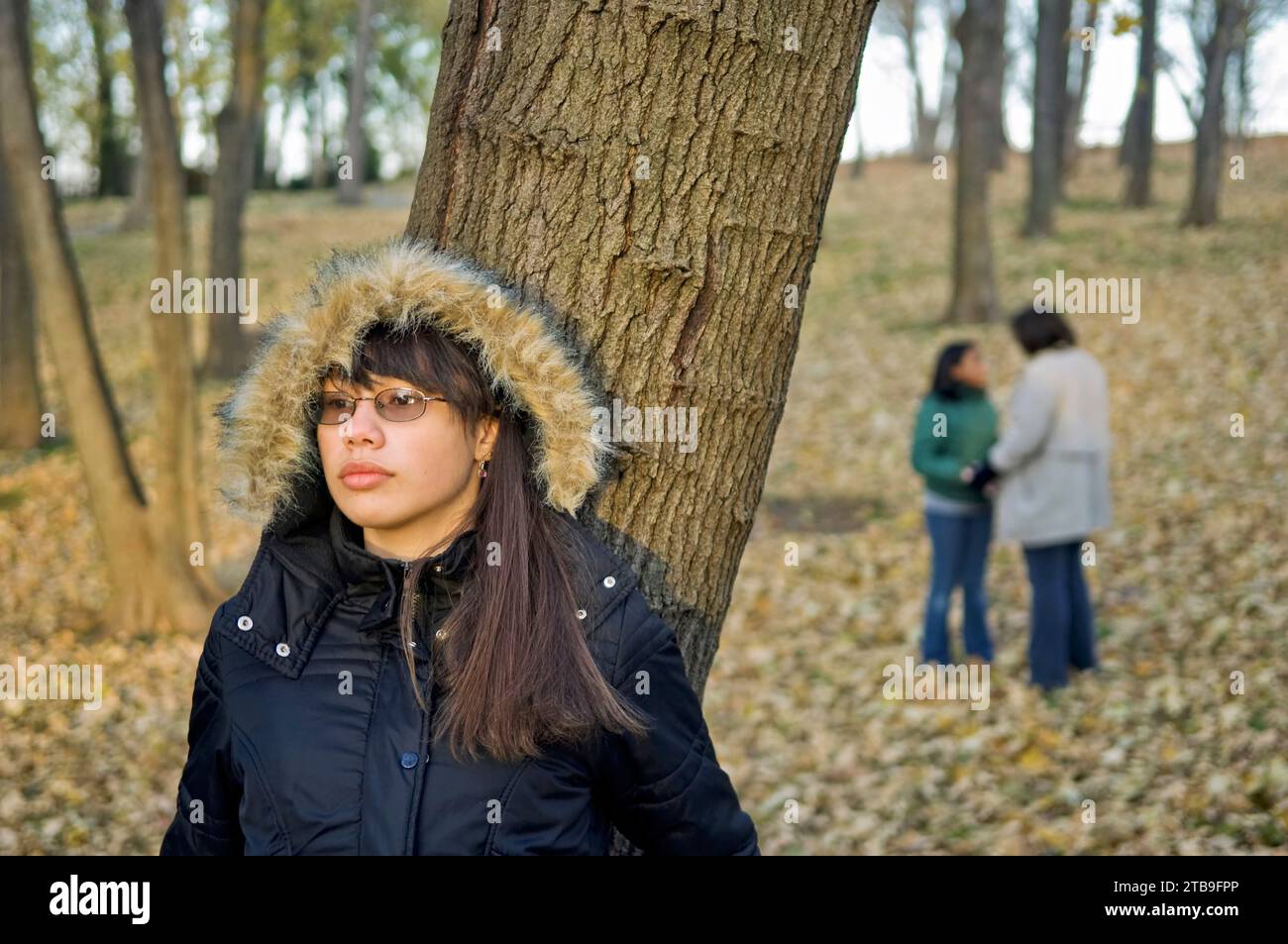 Teenage girl leans against a tree in a city park in autumn, with her mother and sister in the background Stock Photo