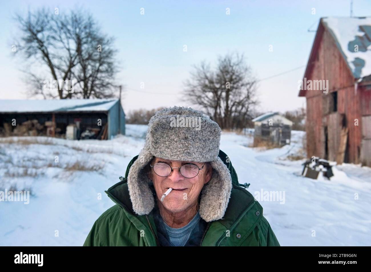 Man smoking a cigarette stands in the snow at a farm; Dunbar, Nebraska, United States of America Stock Photo