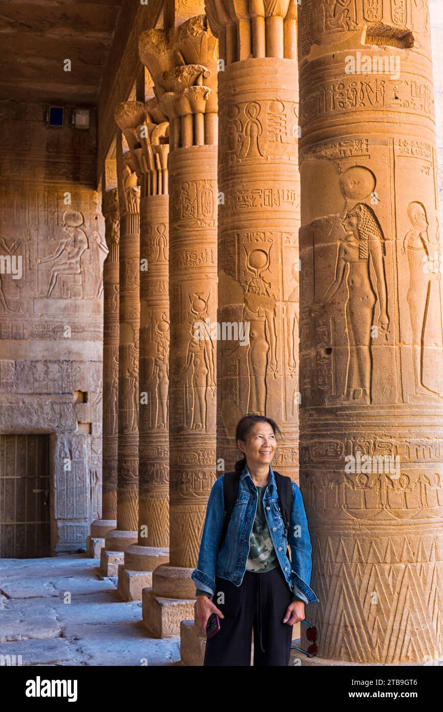 A female Chinese tourist stands among a row of columns with ancient Egyptian hieroglyphic details at the Philae Temple; Aswan, Egypt, Africa Stock Photo
