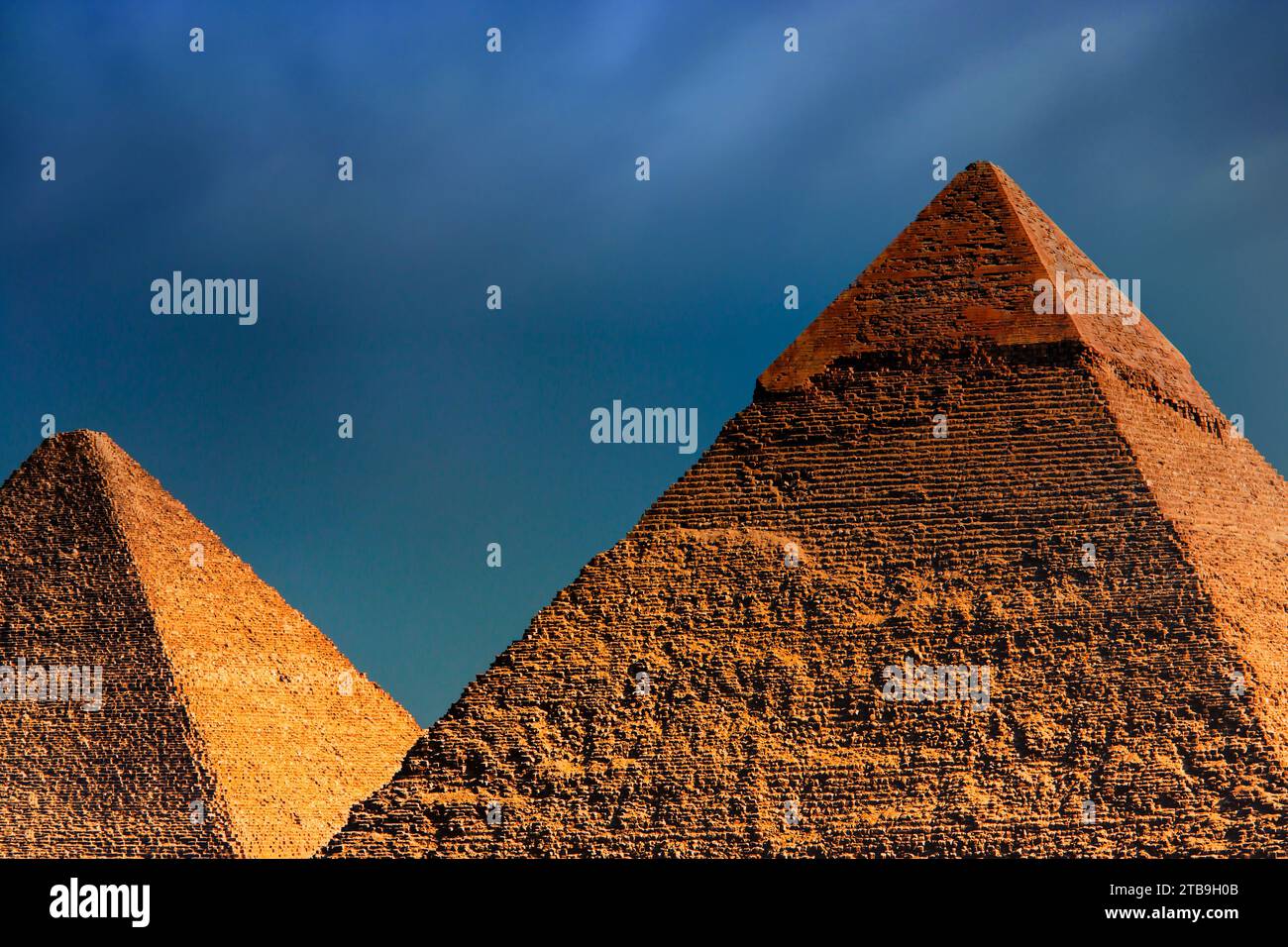 Two of the primary Pyramids of Giza, sunlit with a golden hue, the Great Pyramid (Khufu) under a dark, cloudy sky; Giza, Egypt Stock Photo