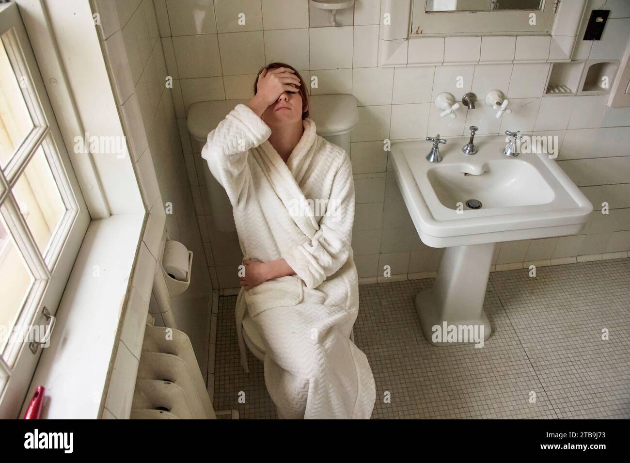 Young woman sits in a bathrobe holding her head in her bathroom; Lincoln, Nebraska, United States of America Stock Photo