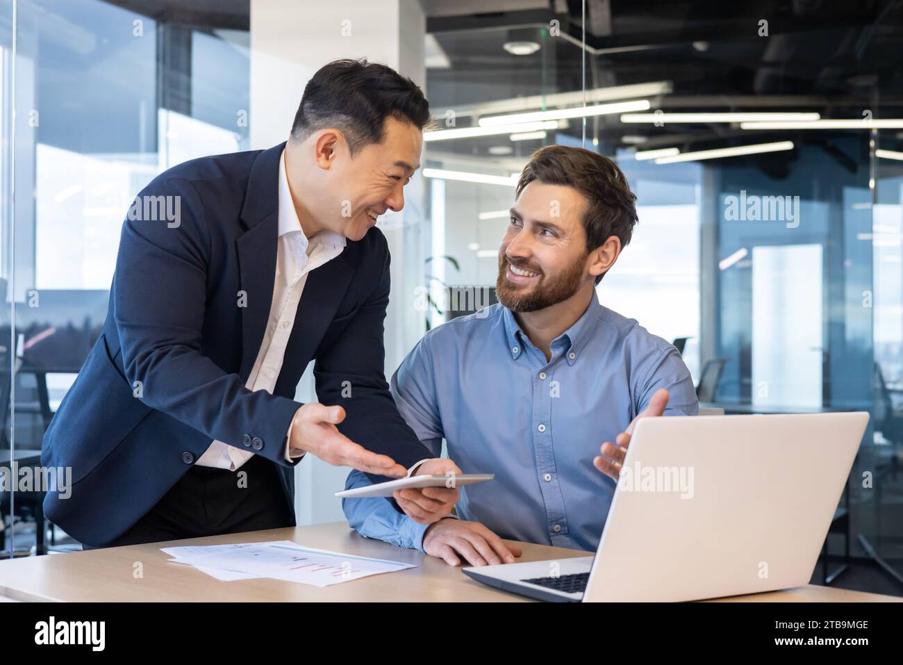 Young two interracial businessmen working in office with documents, agreement, project on tablet and laptop. Stock Photo