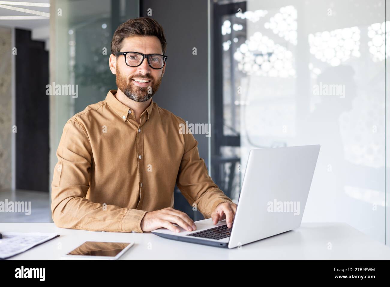 Portrait of successful man at workplace inside office, experienced smiling businessman in shirt smiling and looking at camera, programmer developing new software with laptop. Stock Photo