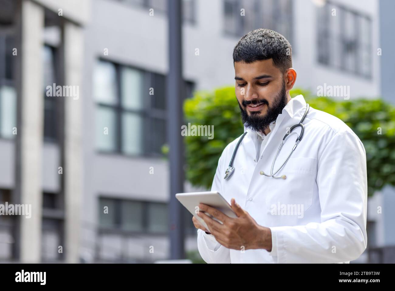 A young Muslim doctor in a bedlam robe stands outside the hospital and uses a tablet, writes, types, consults. Stock Photo