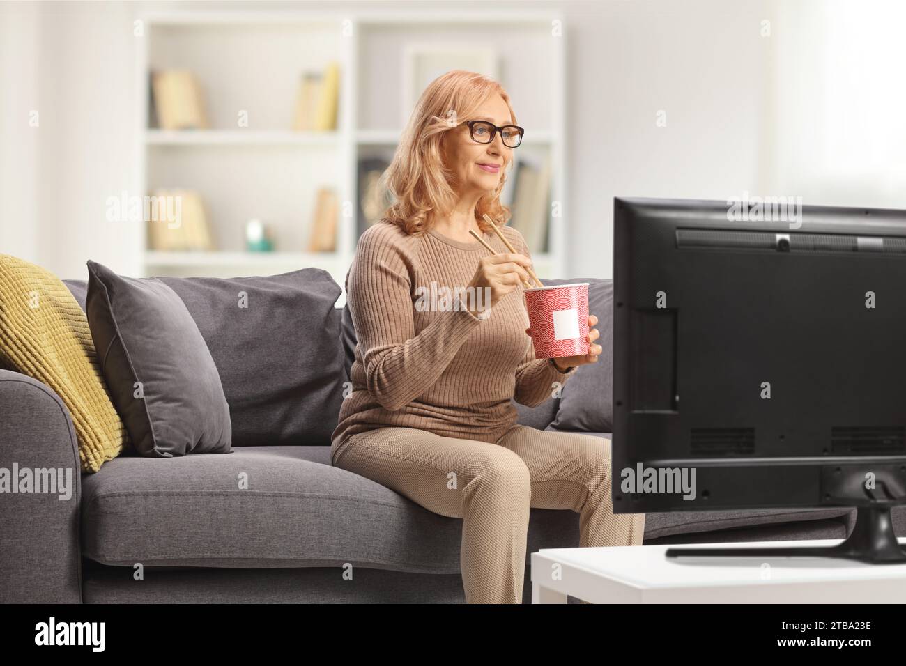 Woman sitting on a couch in front of tv and eating noodles Stock Photo