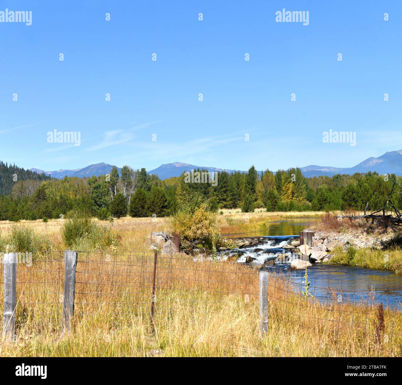 Fence runs in front of a small country stream in Oregon.  Cascades rise in background.  Water splashes over rocks in a small waterfall. Stock Photo