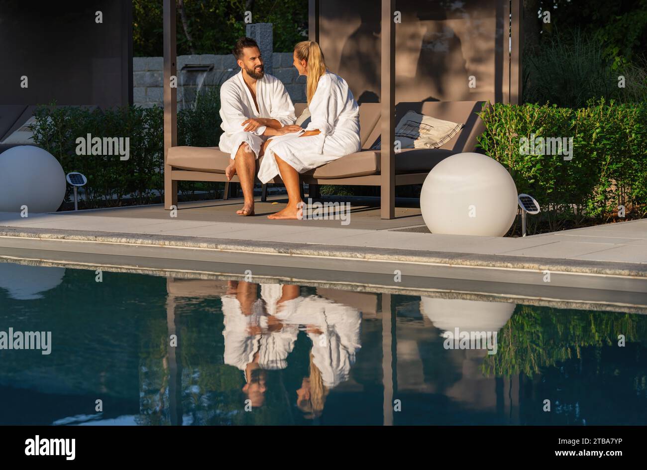 Couple in bathrobes sitting on bench lounger by a pool, reflection in