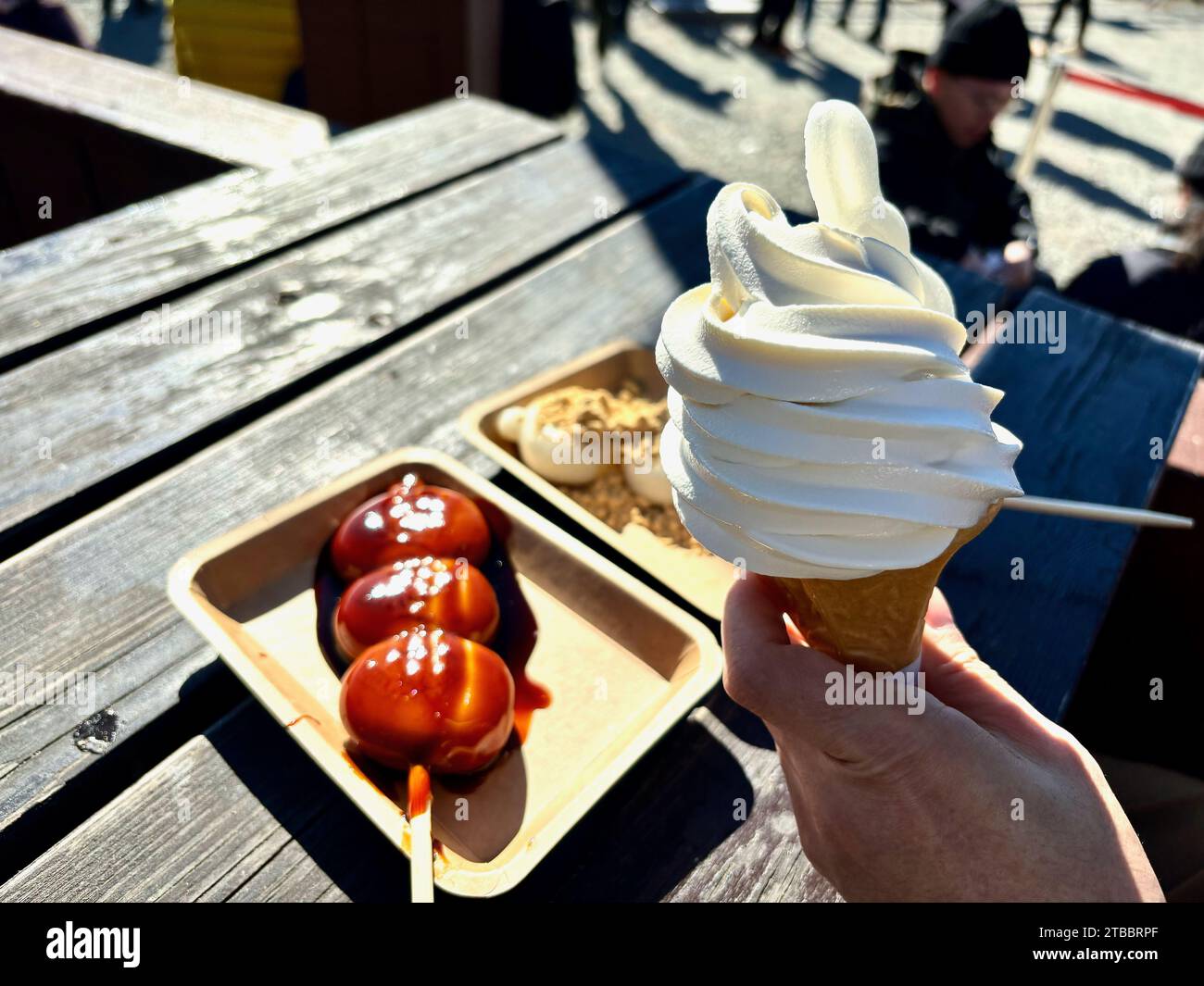 A hand holding an ice cream cone with two plates of Japanese rice dumplings or dango in the background. Stock Photo