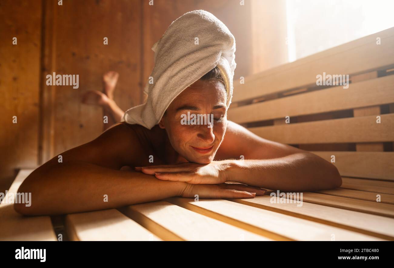 Contented woman lying on a finnish sauna bench with a towel wrapped around her head. Spa wellness hotel concept image. Stock Photo