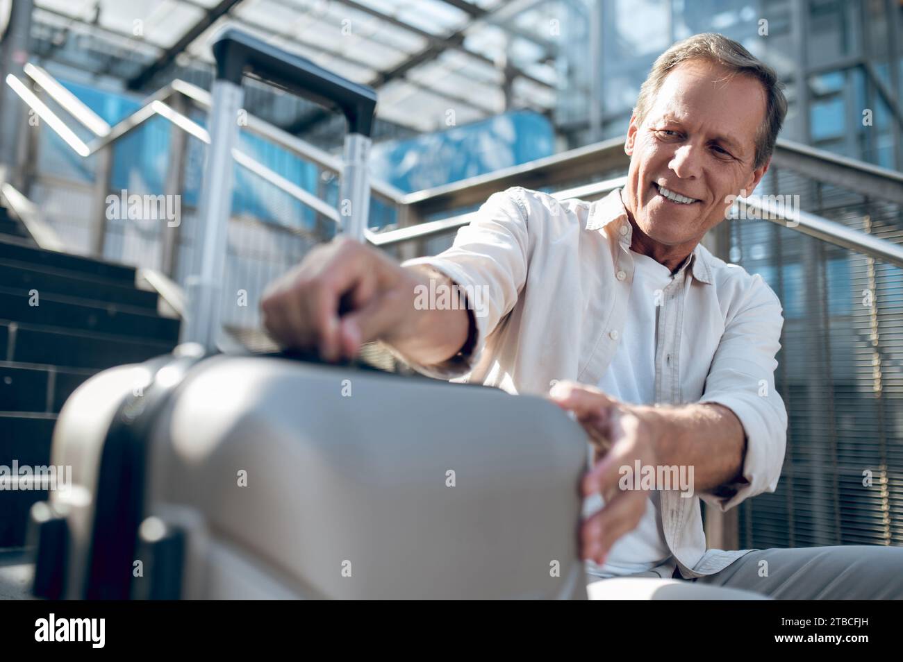 Smiling middle aged man in shirt sitting in street opening his suitcase. Stock Photo