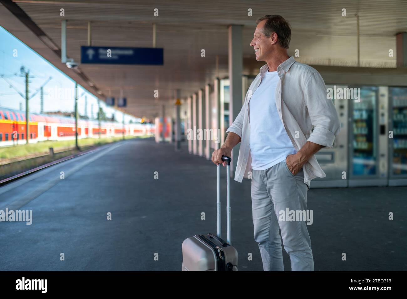 Happy middle aged man with suitcase standing on platform of train station. Stock Photo