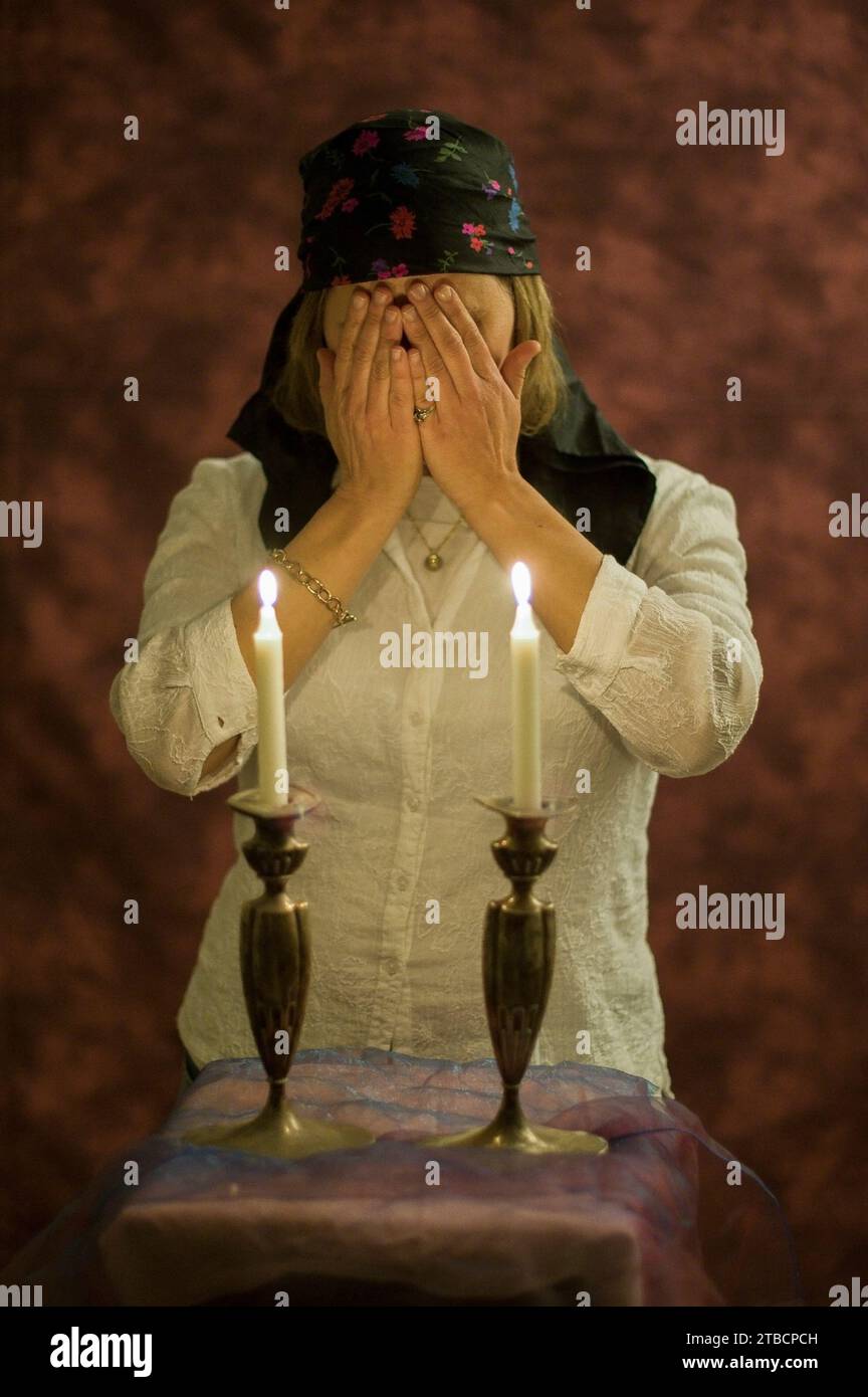 A traditional Orthodox Jewish woman with hair covered by a scarf covers her face with her hands as she recites the blessing over the candles on the ev Stock Photo