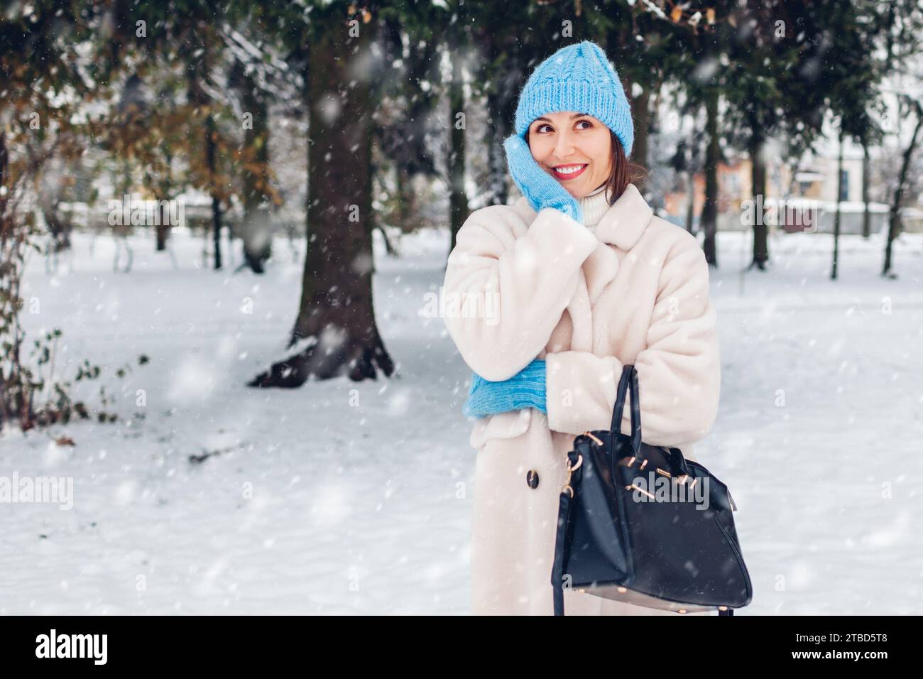 Happy smiling woman walking in snowy winter park wearing white sheepskin fur coat holding handbag enjoying landscape Stock Photo