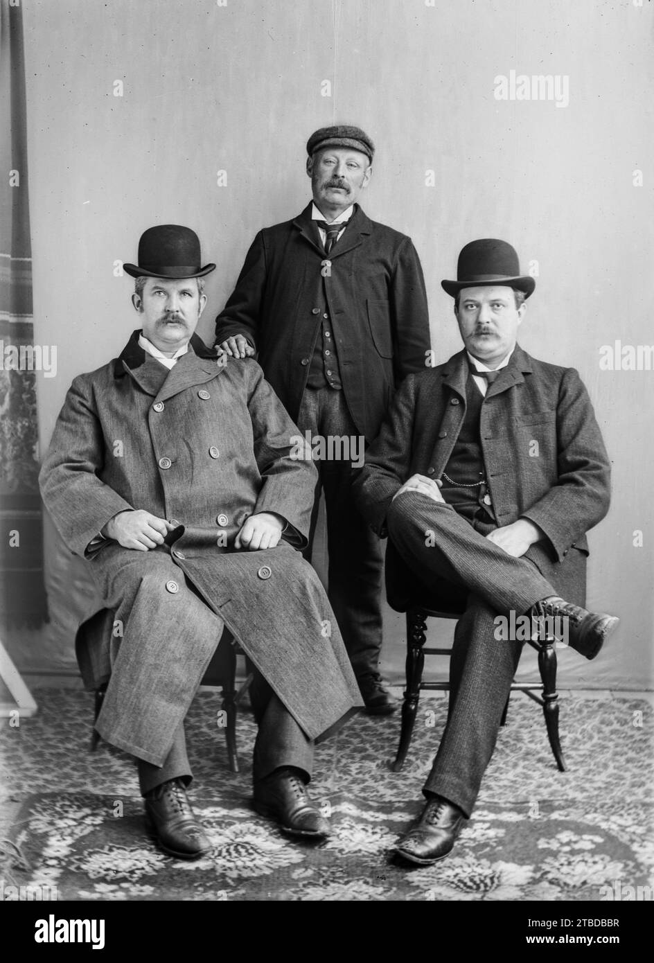 Edwardian formal studio portrait of three men in suits, two sitting and one standing. Bowler hats and a cap.  One in a long coat.  Taken from a vintage glass plate negative.  C1900 – 1910 Stock Photo