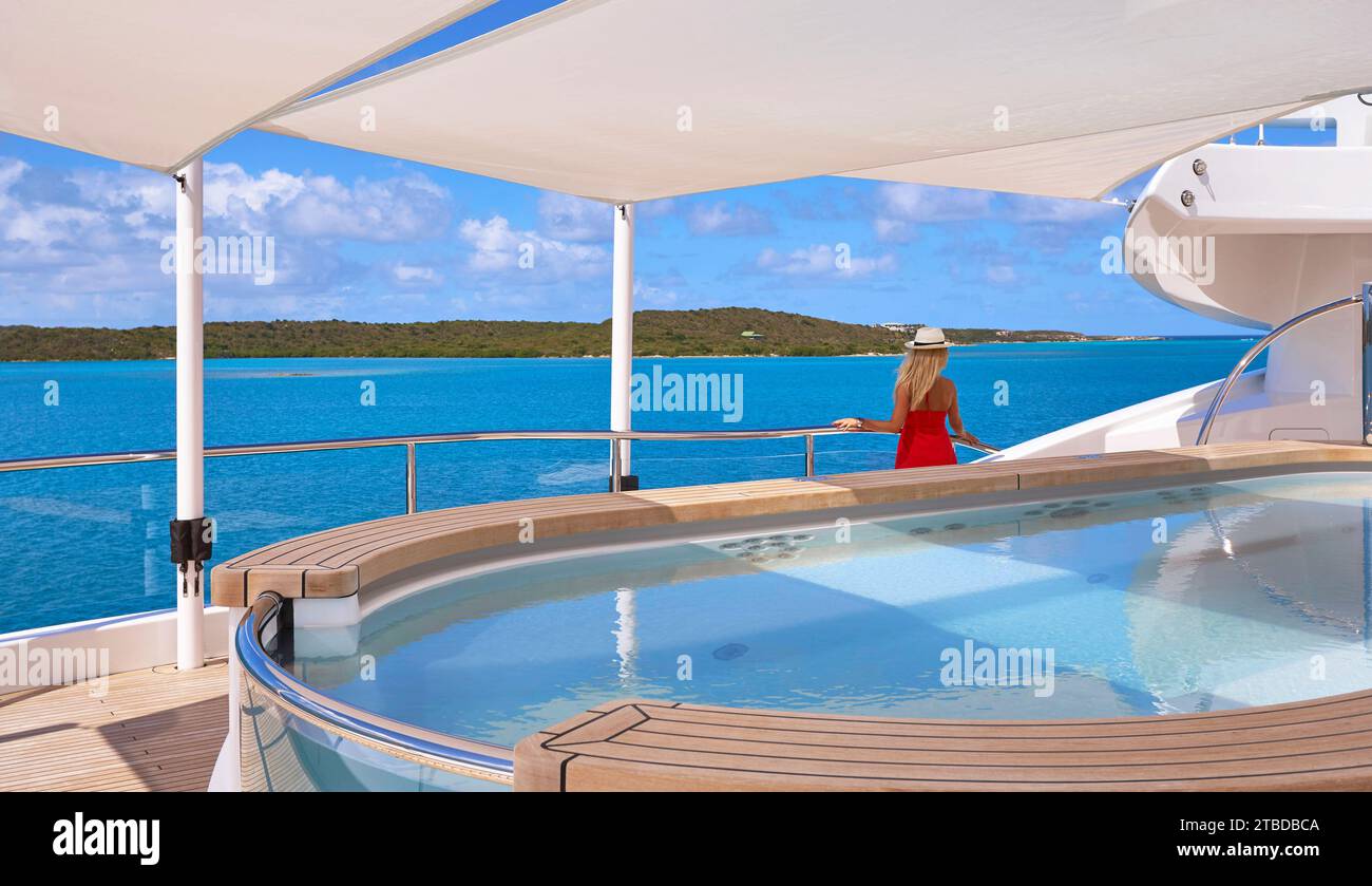 Girl wearing a red dress and a straw hat standing at the rail, with the jacuzzi in the foreground, looking at the turquoise sea and the island beyond. Stock Photo