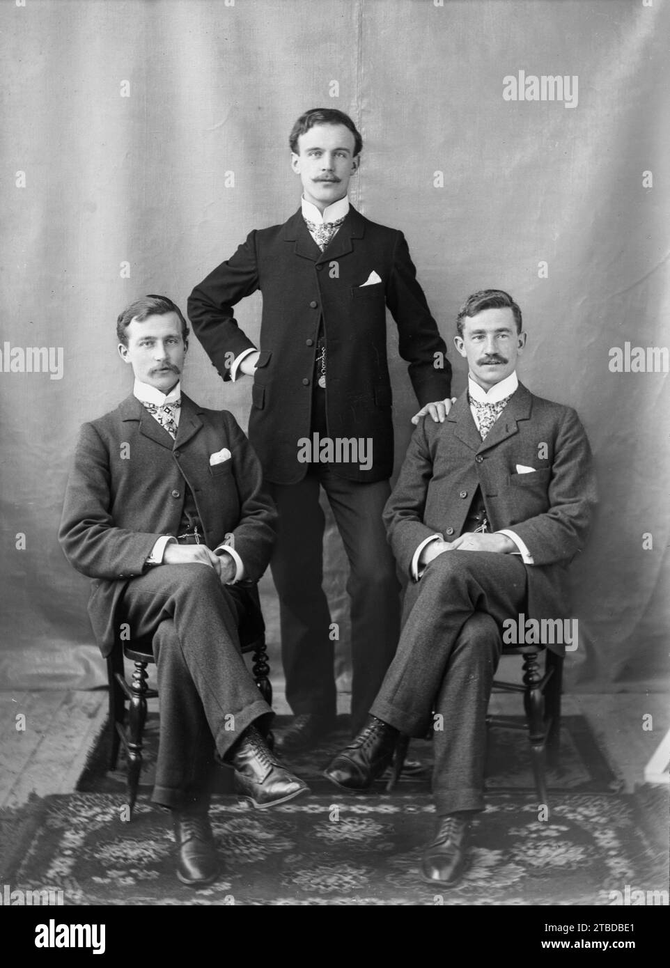 Edwardian formal studio portrait of three men in suits, two sitting and standing. Cravats and stiff collars.  Taken from a vintage glass plate negative.  C1900 – 1910 Stock Photo