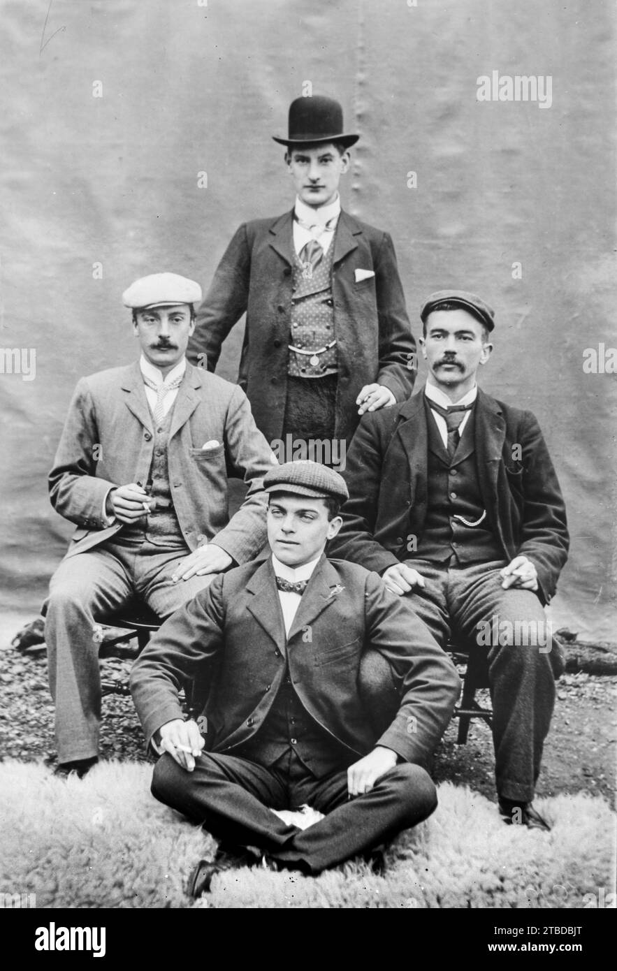 Edwardian formal studio portrait of four men in suits, one standing, one cross legged on a sheepskin rug and two seating on chairs. All smoking.  Taken from a vintage glass plate negative.  C1900 – 1910 Stock Photo