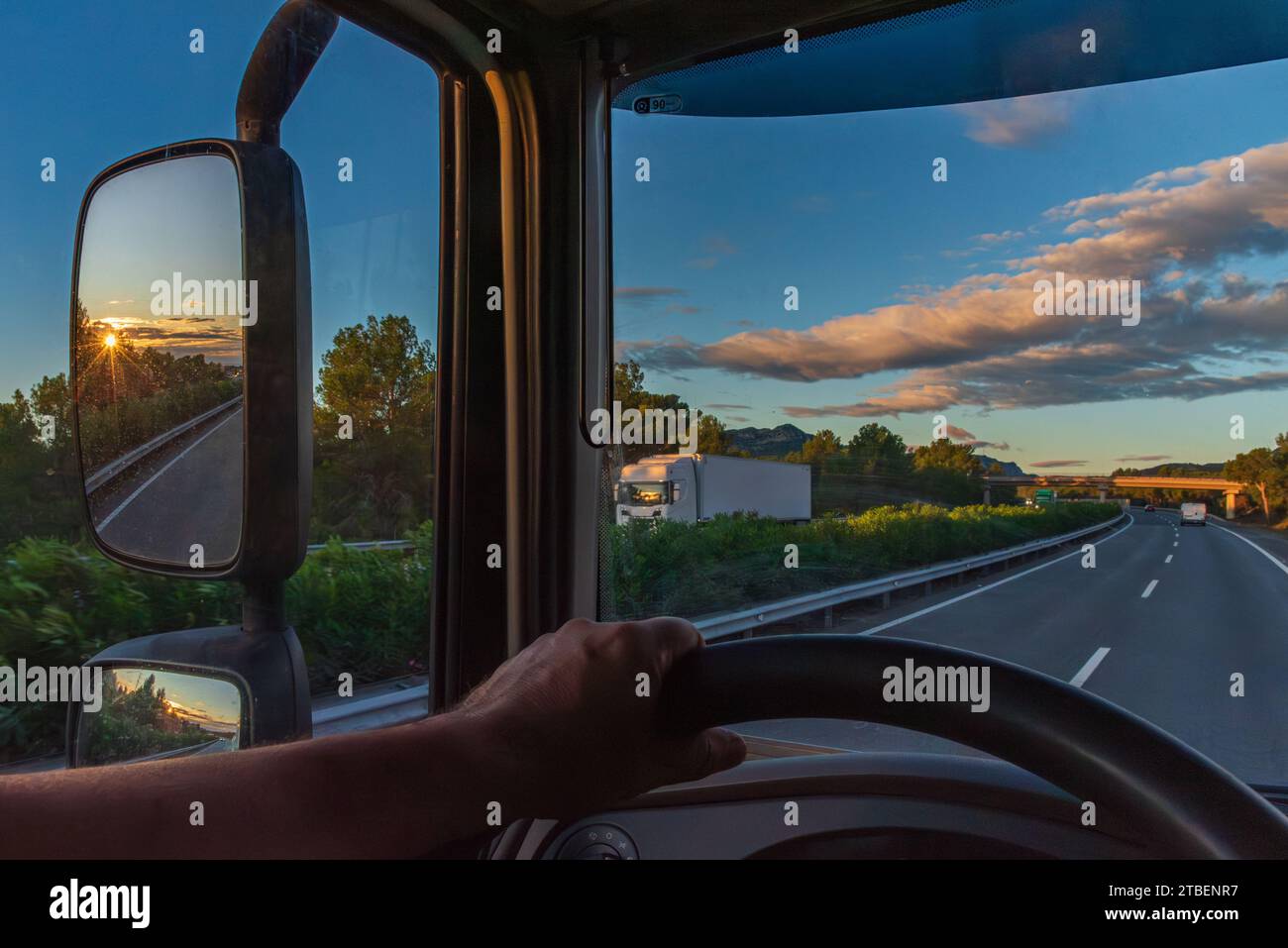 View from the driver's seat of a truck of a highway at dawn and the sun reflected in the rearview mirror. Stock Photo