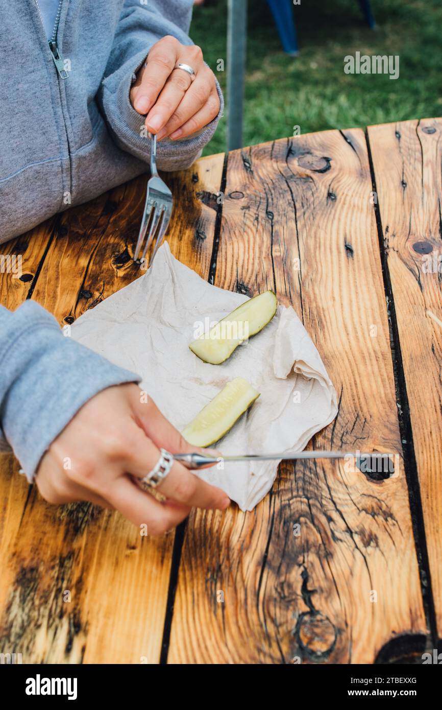 close up of a person holding a knife and fork cutting a pickle spear in half on wooden table outdoors Stock Photo