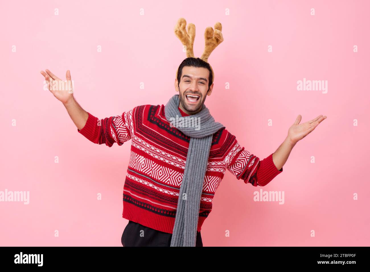Festive studio shot portrait of smiling Caucasian man wearing Christmas sweater and reindeer headband raising hands with palms opening in pink color i Stock Photo