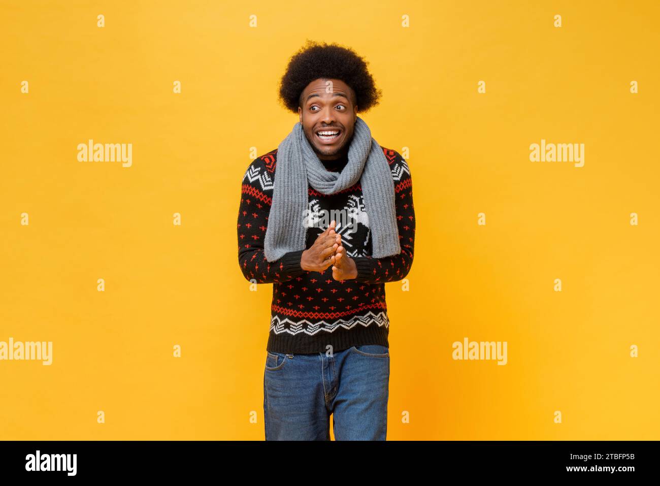 Surprised excited African American man wearing Christmas sweater and scarf, festive studio shot in yellow color isolated background Stock Photo