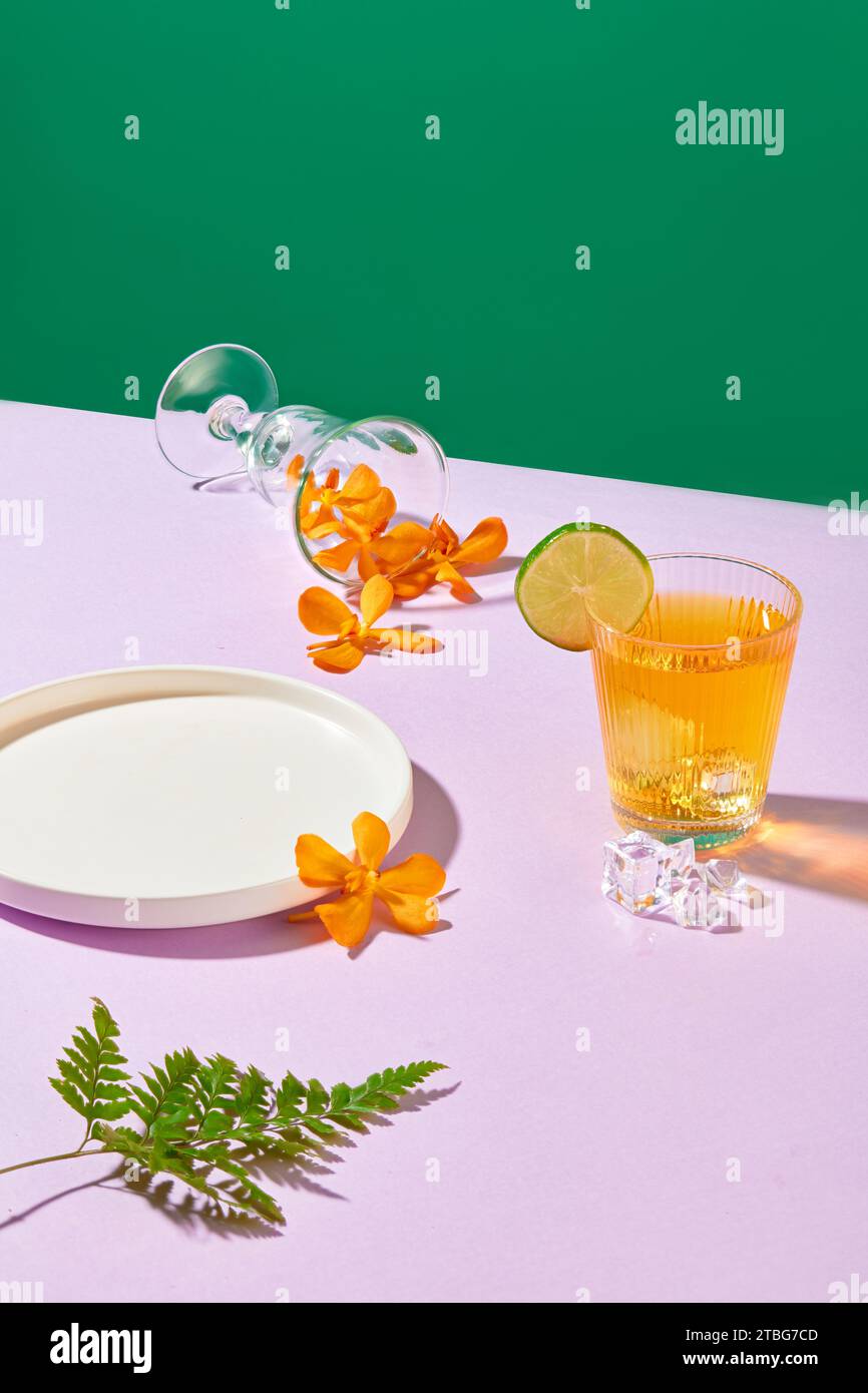 An white plate, a glass containing a transparent yellow liquid decorated with a slice of lemon, a fern leaf, fresh flower petals poured from a cocktai Stock Photo