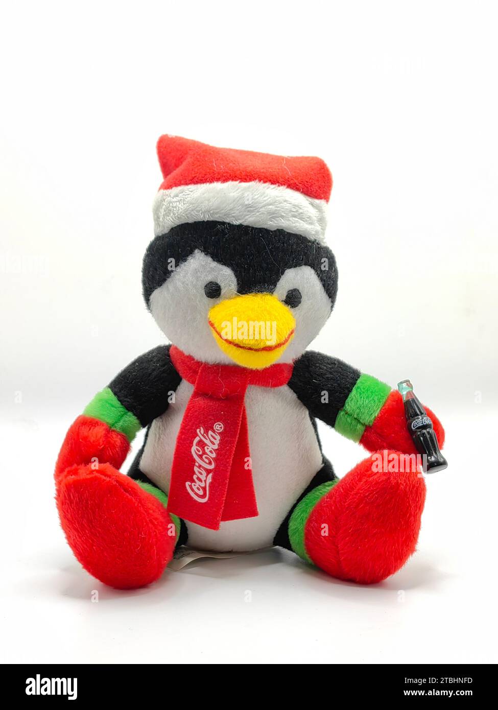 Isolated dark blue penguin,on white paper background. Wearing red christmas hat and scarf with coca cola logo on it.  Toy plush animal.Happy holidays. Stock Photo