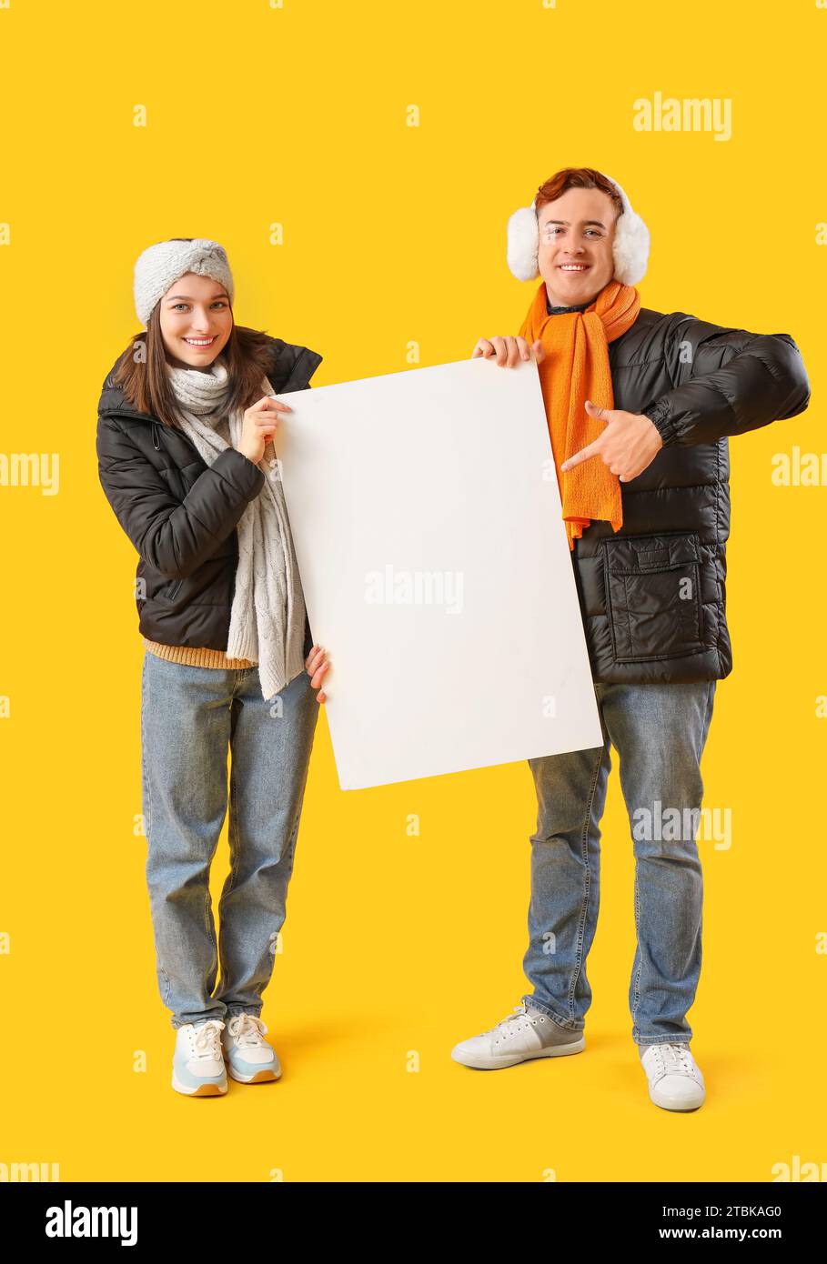 Young couple in winter clothes with blank poster on yellow background Stock Photo