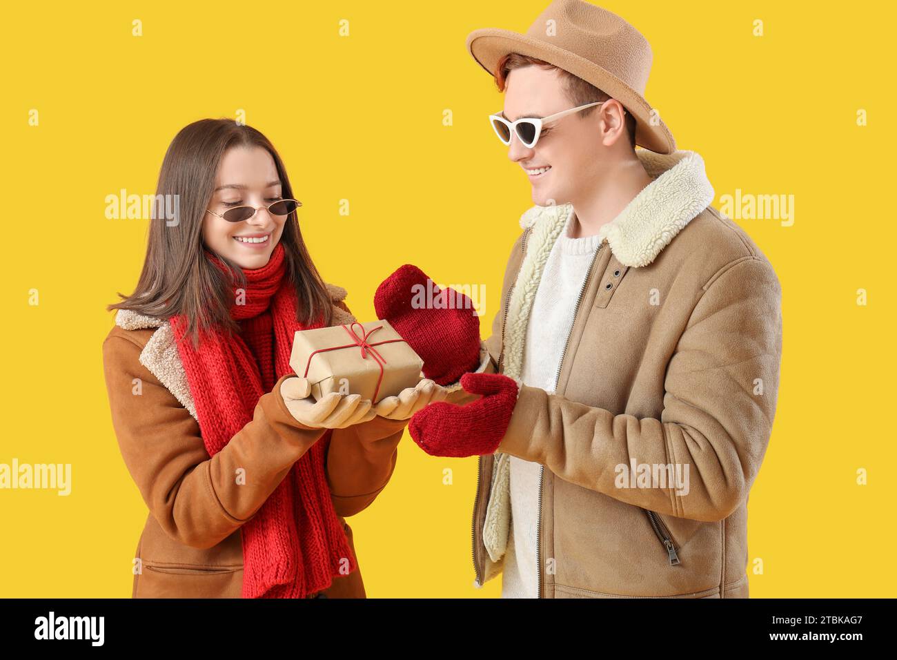 Young couple in winter clothes with Christmas gift on yellow background Stock Photo