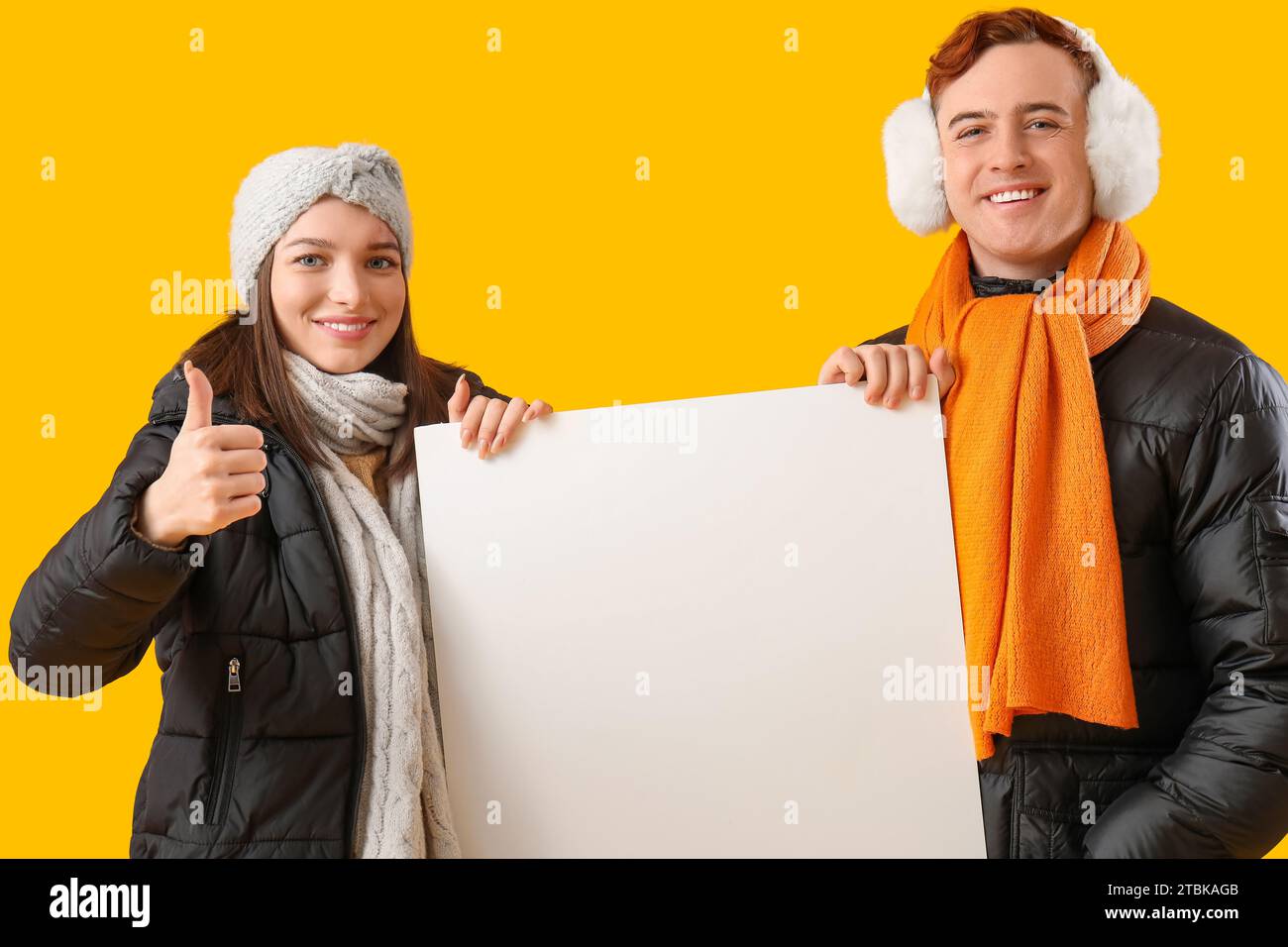 Young couple in winter clothes with blank poster on yellow background Stock Photo