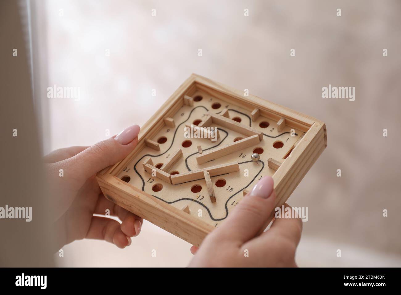 Woman holding wooden toy maze with metal ball on blurred background, closeup Stock Photo