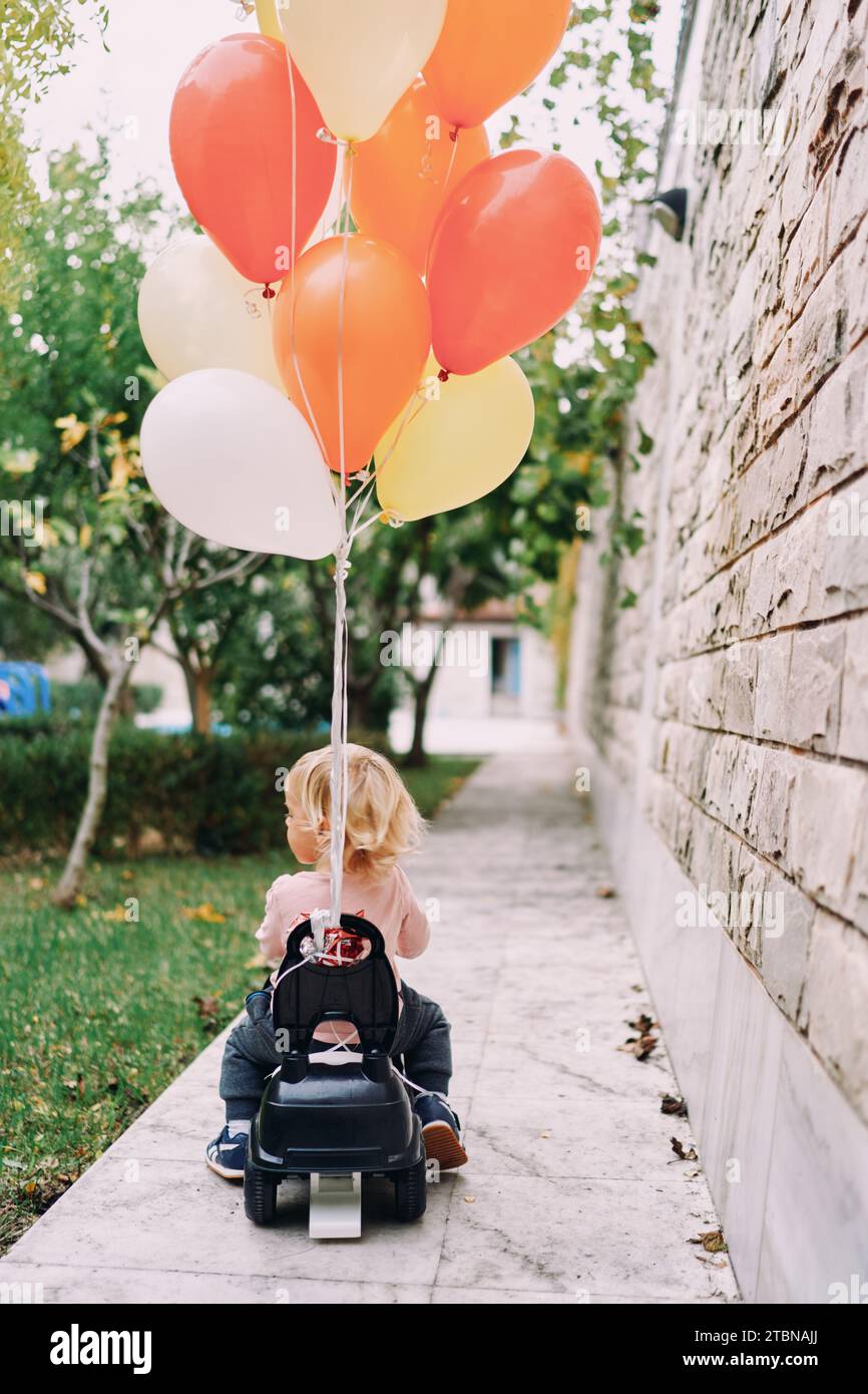 Little girl is driving a toy car with tied balloons around the yard ...