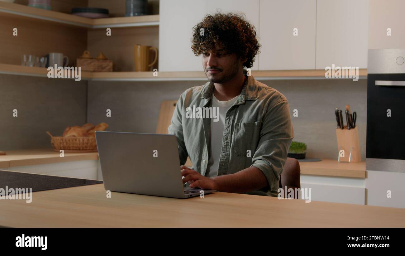 Concentrated Indian businessman typing computer working shopping online distant from home with laptop at kitchen serious focused freelancer Arabian Stock Photo
