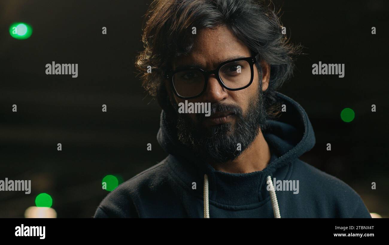 Male portrait serious confident bearded Indian 30s millennial business man in eyeglasses looking to side turn head at camera at dark parking lot Stock Photo