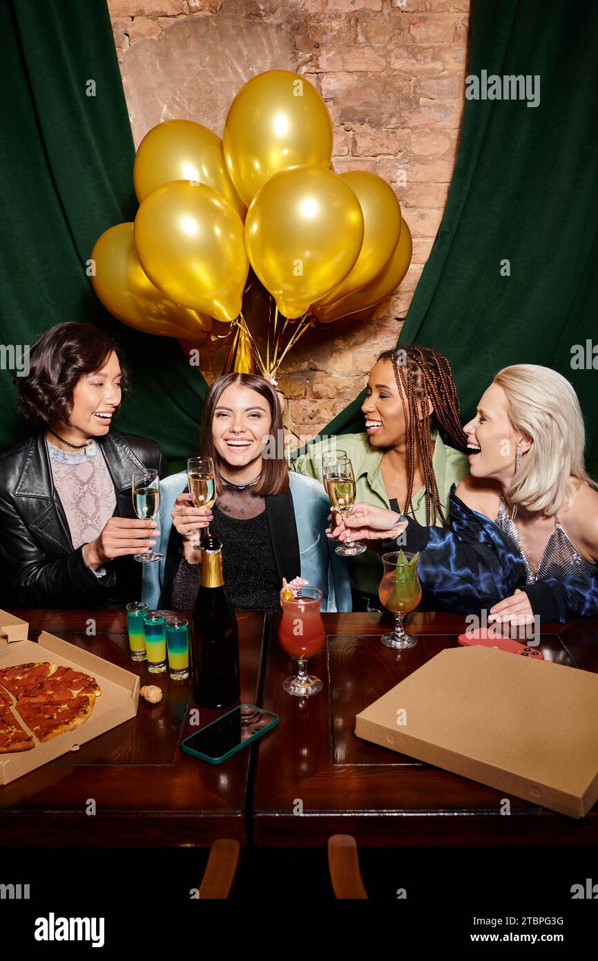 cheerful multiracial fashionistas with champagne near cocktails and golden balloons in bar, birthday Stock Photo