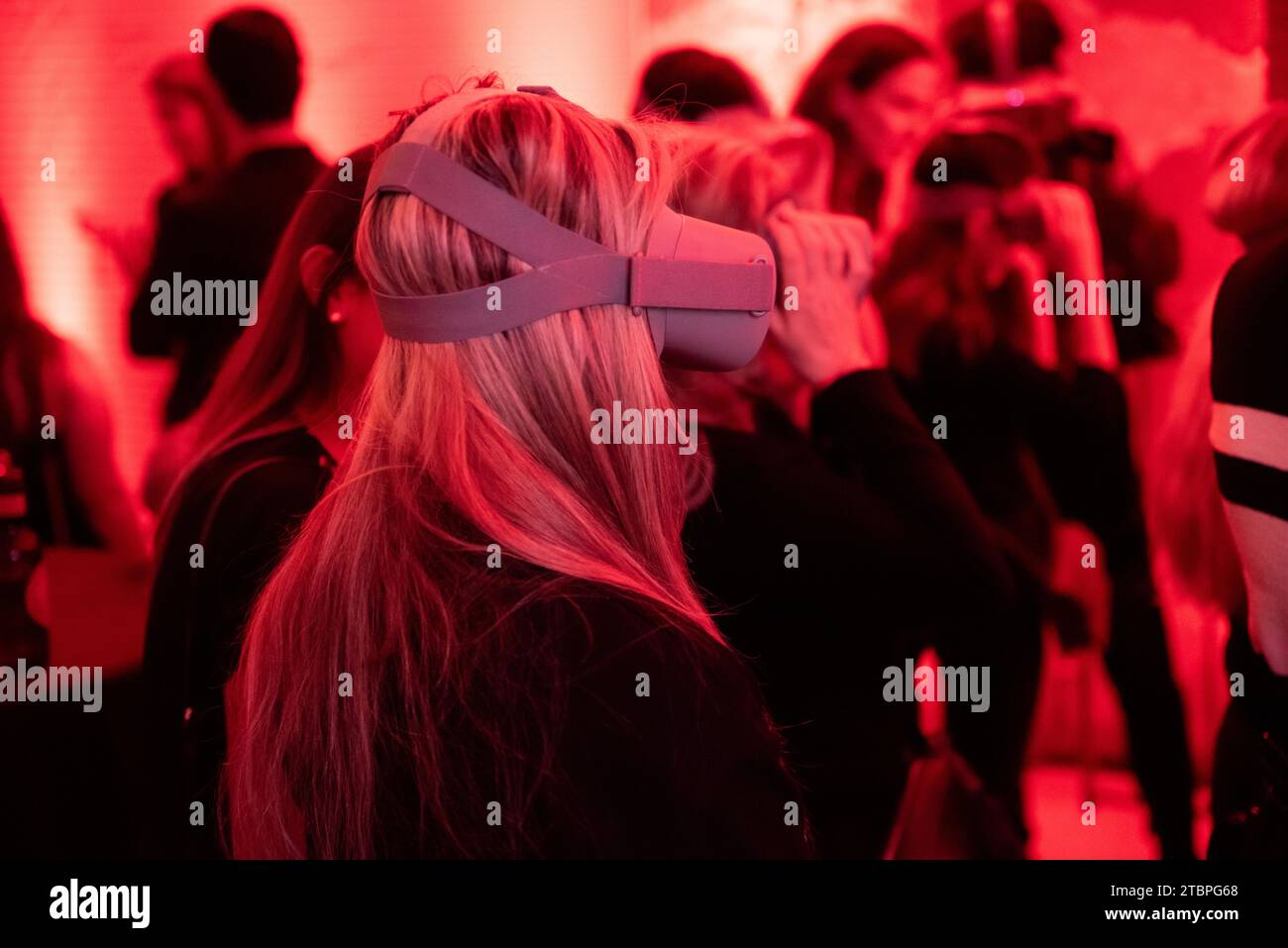 Silhouetted in red radiance, a woman in profile navigates a virtual realm, surrounded by fellow enthusiasts, in a symphony of shared experiences at a Stock Photo