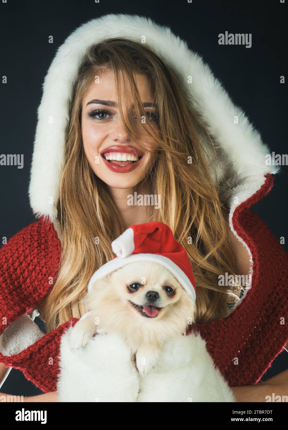 Smiling Santa woman in fur hat and white gloves with Pomeranian Spitz. Christmas girl with small dog in Santa Claus hat. Holiday pet market. Animals Stock Photo