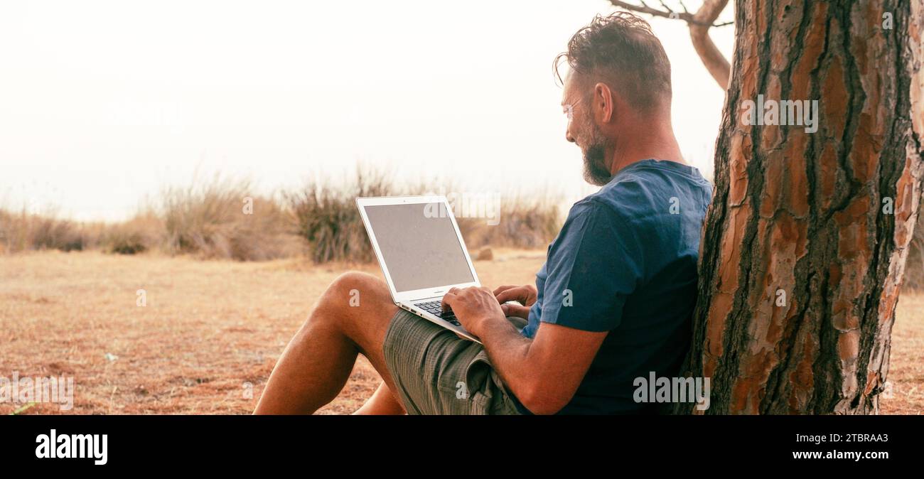 One modern traveler free worker freelance lifestyle working on laptop sitting on the ground against a tree and enjoying nature. Outside computer connection and small online business job technology. Stock Photo