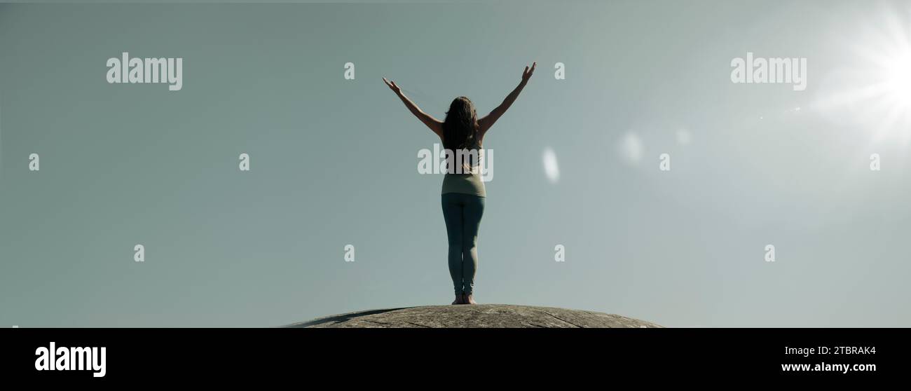 A woman stands silhouetted on a rock with arms outstretched Stock Photo