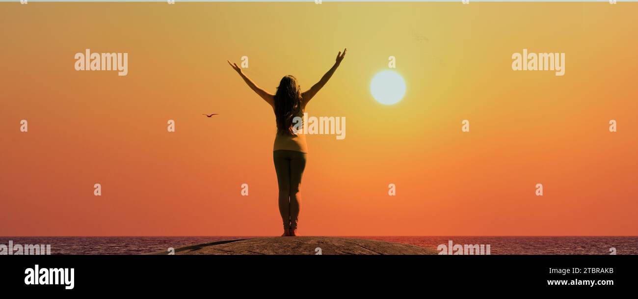 A woman stands silhouetted on a rock by the sea with her arms outstretched Stock Photo
