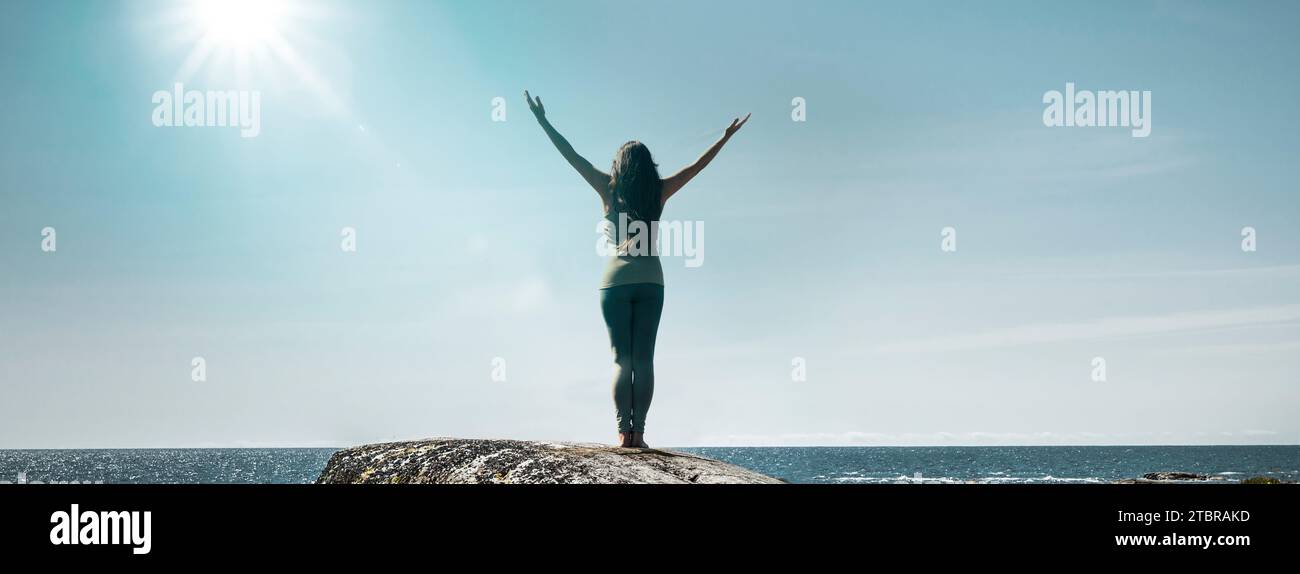 A woman stands silhouetted on a rock by the sea with her arms outstretched Stock Photo