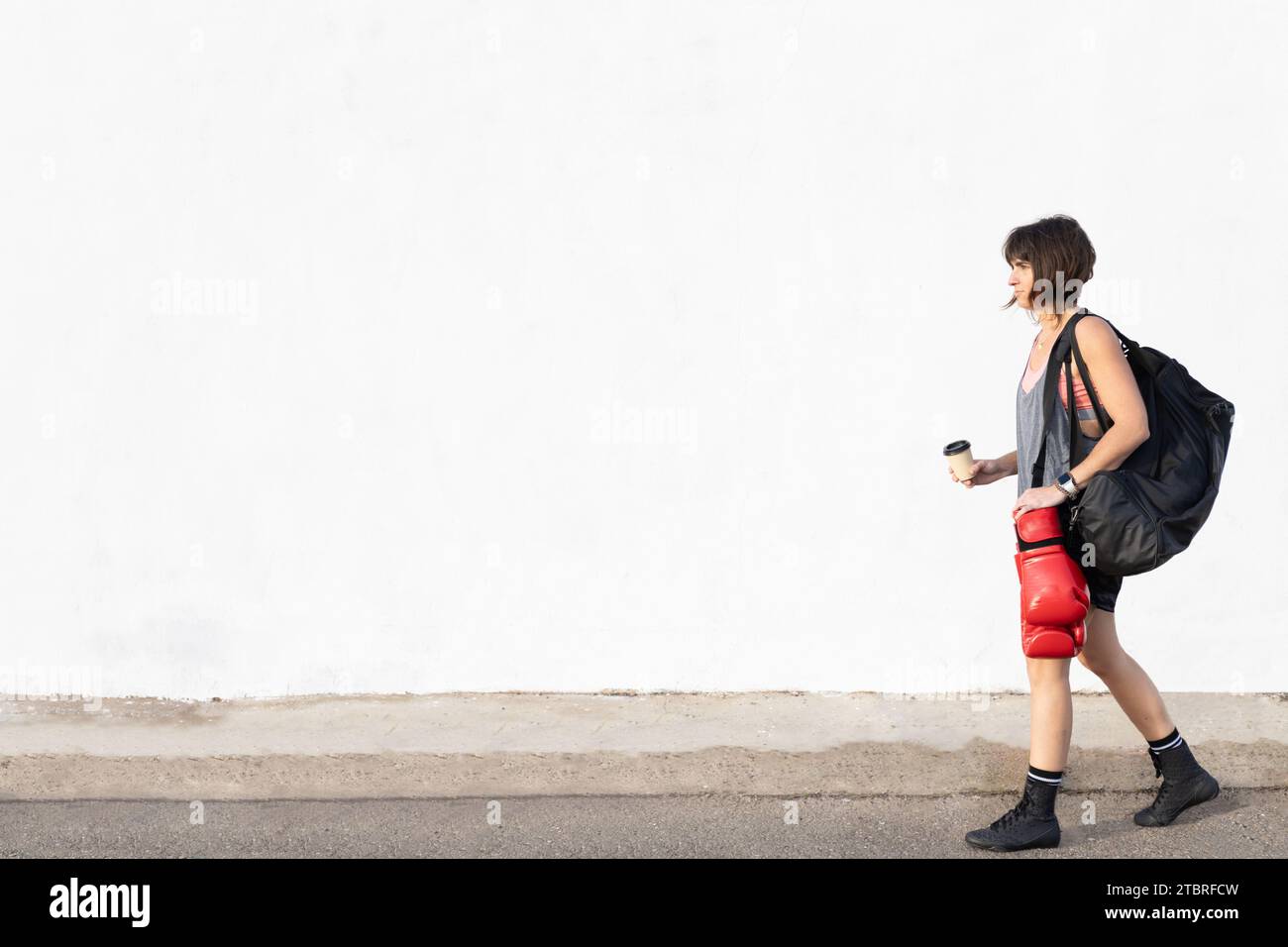 Horizontal photo woman mid adult Caucasian walking to gym with sports clothes, a backpack, red boxing gloves and a coffee to carry in hand. Copy space Stock Photo