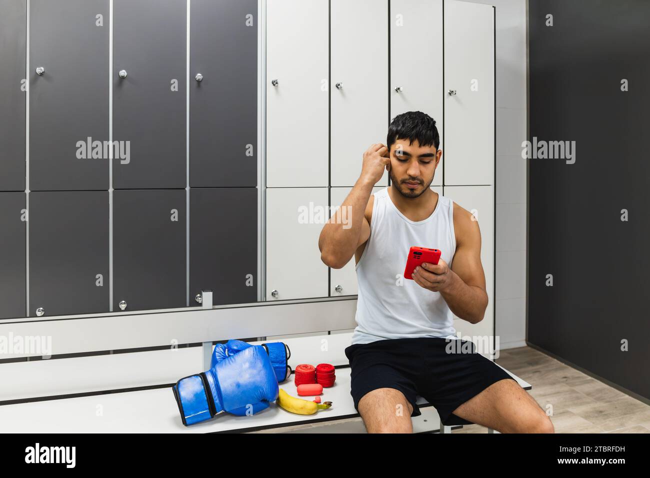 Horizontal photo man young adult mixed race sitting in the van of the boxing gym locker room with his cell phone in his hand and placing headphones in Stock Photo