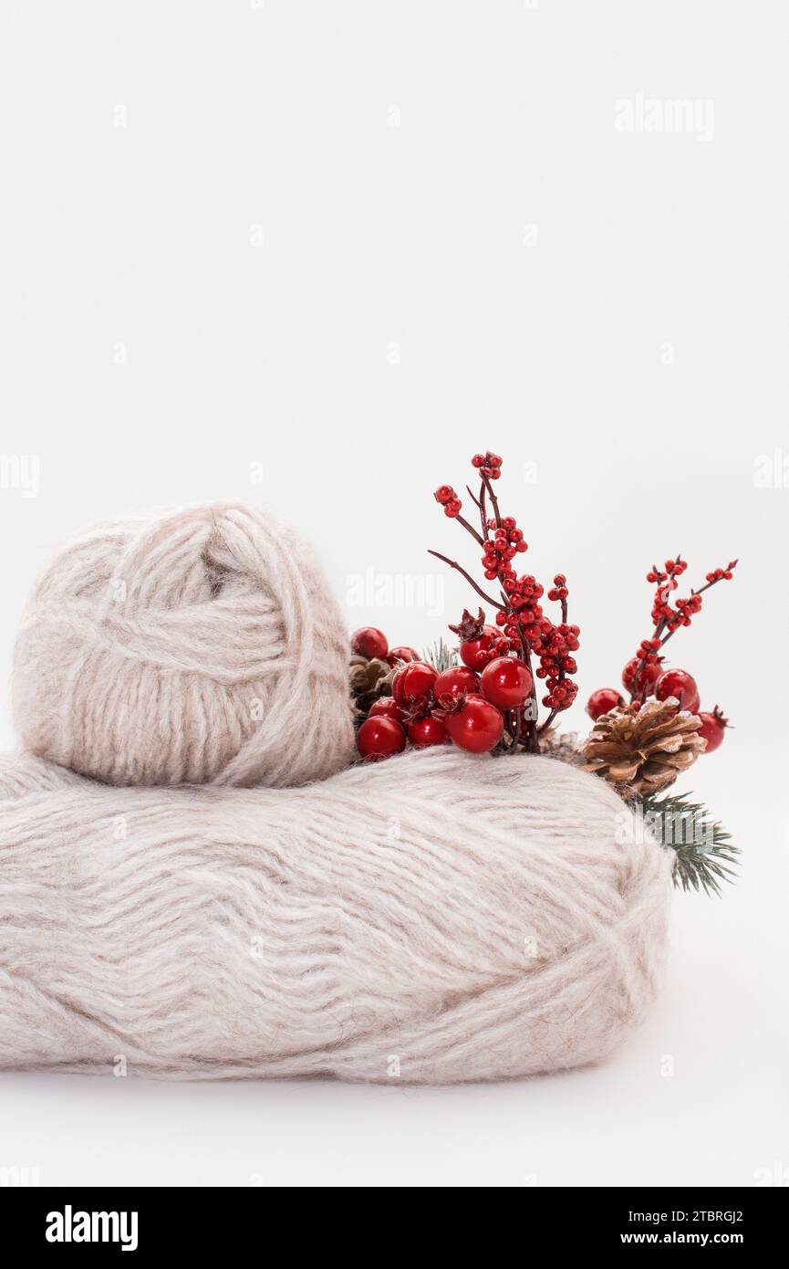 Gray wool skeins or wool yarn with branch of red berries and pine cones on white background. Handmade, knitting, hobby. Copy space. Vertical. Side vie Stock Photo