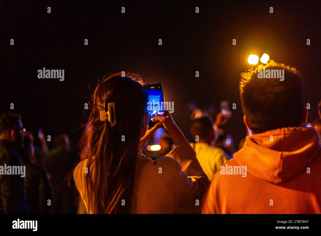 An anonymous young woman recording a light show on her phone at night, seen from behind, live streaming from her smartphone Stock Photo