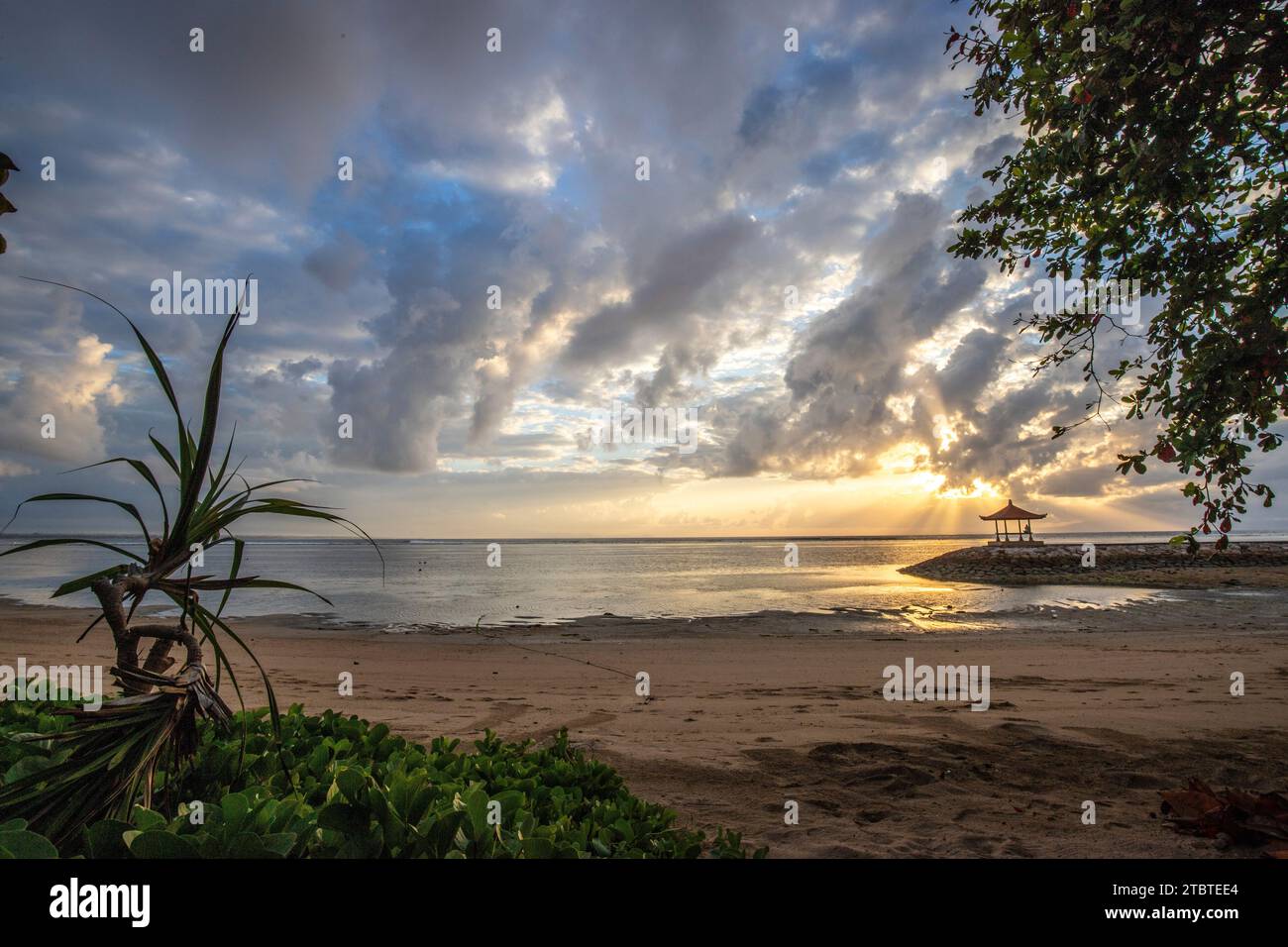 Sunrise over the sea, view from the sandy beach to the horizon, in the sea there are breakwaters with small temples, calm water with small waves and reflections on the tropical beach of Sanur, Bali, Indonesia Stock Photo
