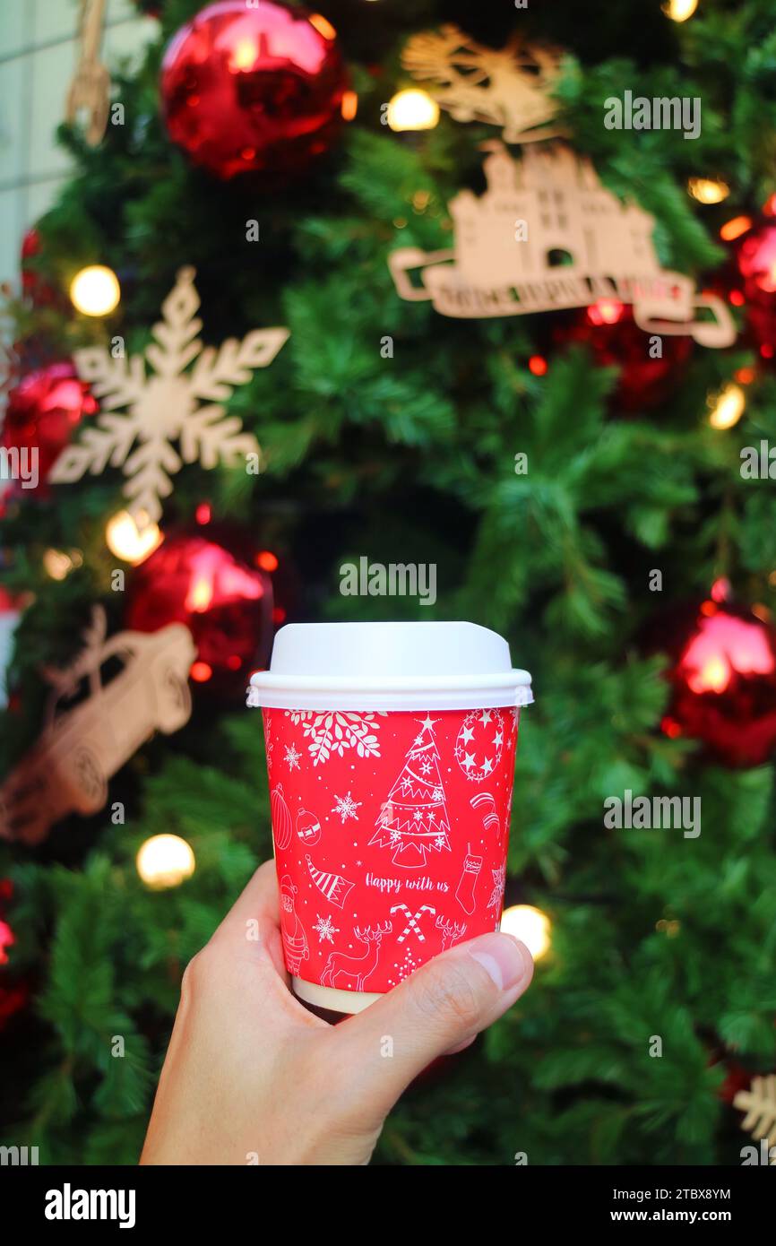 Hand Holding a Holiday Theme Takeaway Coffee Cup with Blurry Christmas Tree in Background Stock Photo