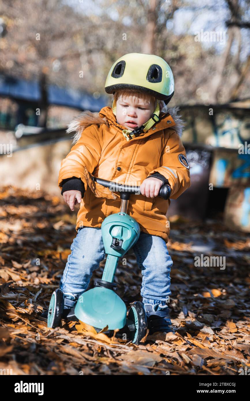 Adorable toddler boy wearing yellow protective helmet riding baby scooter outdoors on autumn day. Kid training balance on mini bike in city park. Fun autumn outdoor activity for small kids Stock Photo