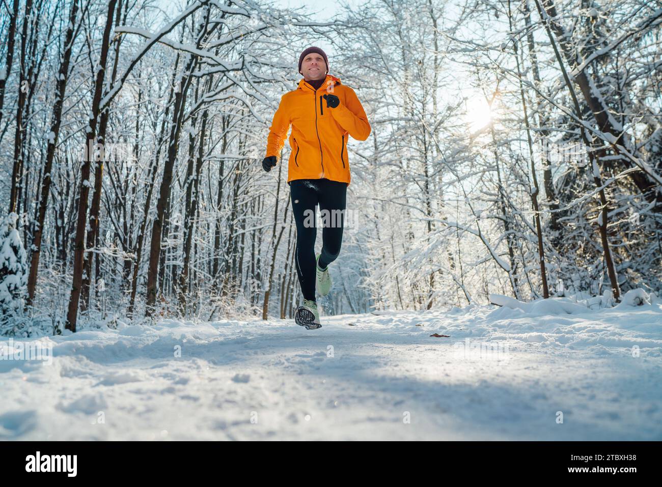 Middle-aged smiling trail runner man dressed bright orange windproof jacket endurance fast running picturesque snowy forest during sunny frosty day. S Stock Photo
