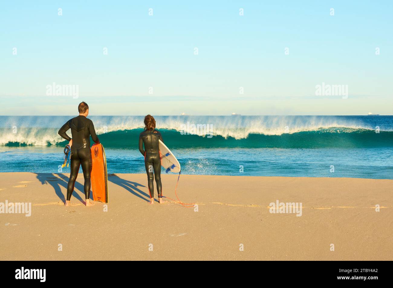 Man with a bodyboard and boy with a surfboard watching a wave breaking in early morning light at Trigg Beach, Perth, Western Australia. Stock Photo